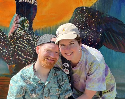 In front of a colorful mural of a loon taking flight, a young white man with a backwards ball cap, red beard, and Hawaiian shirt and a white woman with brown hair and a tie-dye shirt. Both are smiling at the camera