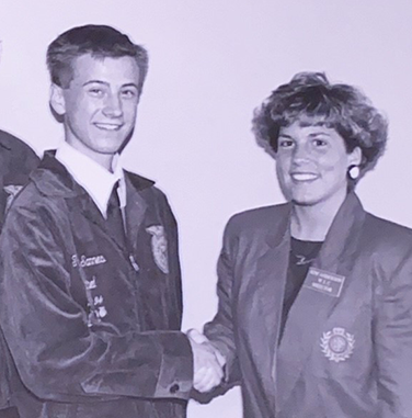 A black and white photo of teenaged Trevor Sannes, a tall young white man, wearing an FFA jacket and shaking hands with a young white woman with curly hair