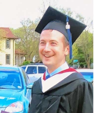 Trevor in a parking lot smiling in the sunshine, wearing his college graduation cap and gown