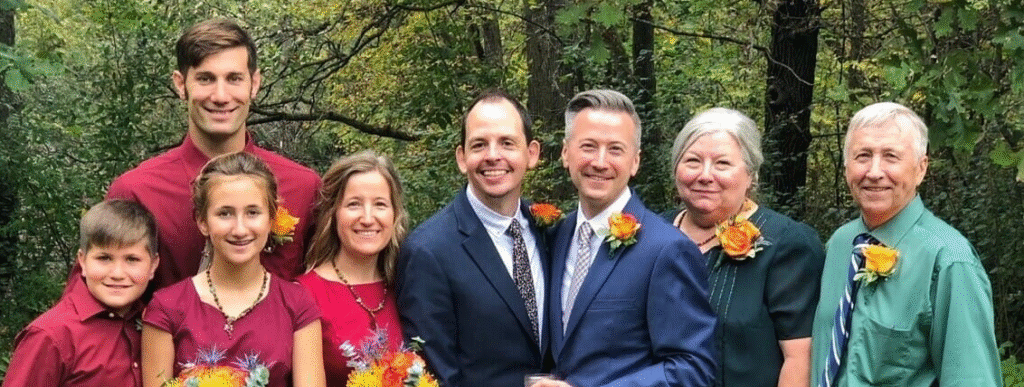 Trevor at his wedding, posing in a beautiful green foresty area with his husband, surrounded by other family members of all ages, wearing or carrying orange roses