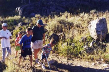 A photograph from the 1990s showing a mom and four boys hiking through a grassy area with large rocks in the background. One of the boys is using forearm crutches.