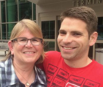 Betsy, a white woman with glasses and shoulder-length dark blonde hair, stands with her son, a young man with short brown hair. Both are smiling at the camera
