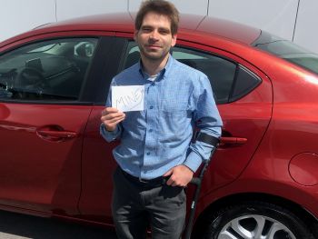 A young white man with brown hair and a forearm crutch stands in front of a red car, holding a card that says "Mine"