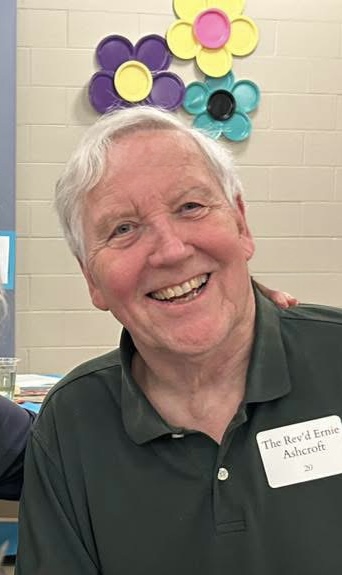 A white man in his 70s with white hair, smiling warmly at the camera. He is wearing a polo shirt with a nametag that says "The Rev'd Ernie Ashcroft"