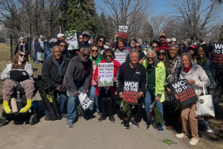 Several dozen people pose for a picture. They are a diverse group of many races, ages, and disabilities. They are carrying palm branches and signs saying things like "Blessed are those who heal the sick" and "woe to the greedy billionaires."
