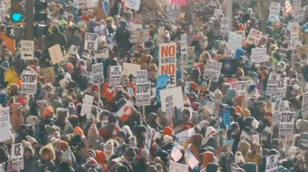 A huge crowd of people, all bundled up for cold weather, fill a downtown street marching and carrying signs protesting ICE.