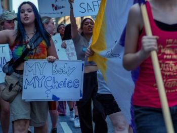 A group of young women march in a protest. One carries a sign reading "My Body, My Choice"
