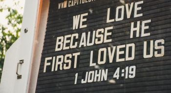 A changeable letterboard sign outside a church reading "We love because he first loved us. 1 John 4:19"