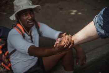 A young male hiker wearing a bucket hat and backpack is sitting on the ground, looking tired and uncertain, reaching out his arm to be helped up by another person outside of the camera fram