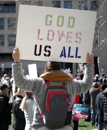 In a crowd gathered in front of a city building, a person holds up a sign reading "God loves us all"