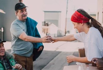 A young woman volunteer wearing a bandana, hands a water bottle to an older man while another older man in a wheelchair looks on