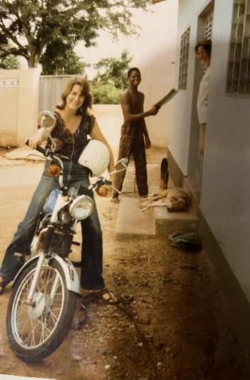 A photo from the 1970s of Judy DesHarnais as a young woman posing on a motorcycle in front of a small concrete building in Benin, while two other young adults smile in the background
