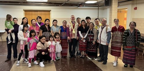 A large group of people in a church basement posing for a photo. Their ages range from toddlers to elderly. Most of the people are Southeast Asian and wearing traditional Karen clothing. The Rev. Judy DesHarnais is in the group, as is Bishop Craig Loya, wearing a traditional Karen sash and necklace.