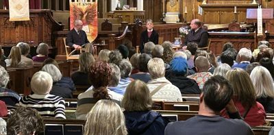 A photo from the rear of St. John's sanctuary, showing many full pews, and at the front Bishop Mariann Budde and Brian Bliss talking with editor Andrew Karre.