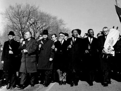 A black-and-white photo showing ministers, rabbis, and other men in suits marching down a street. The Rev. Martin Luther King, Jr. is one of them. Most are carrying small American flags. One rabbi carries the Torah.