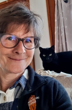 Christine Jacox, who has short brown hair and cat's-eye glasses, takes a selfie with her black cat behind her